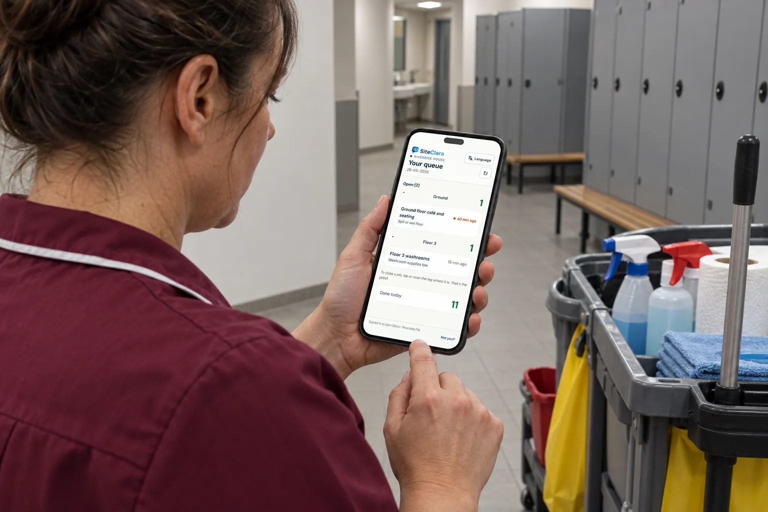 A cleaner checking her job queue on a phone beside a cleaning trolley.