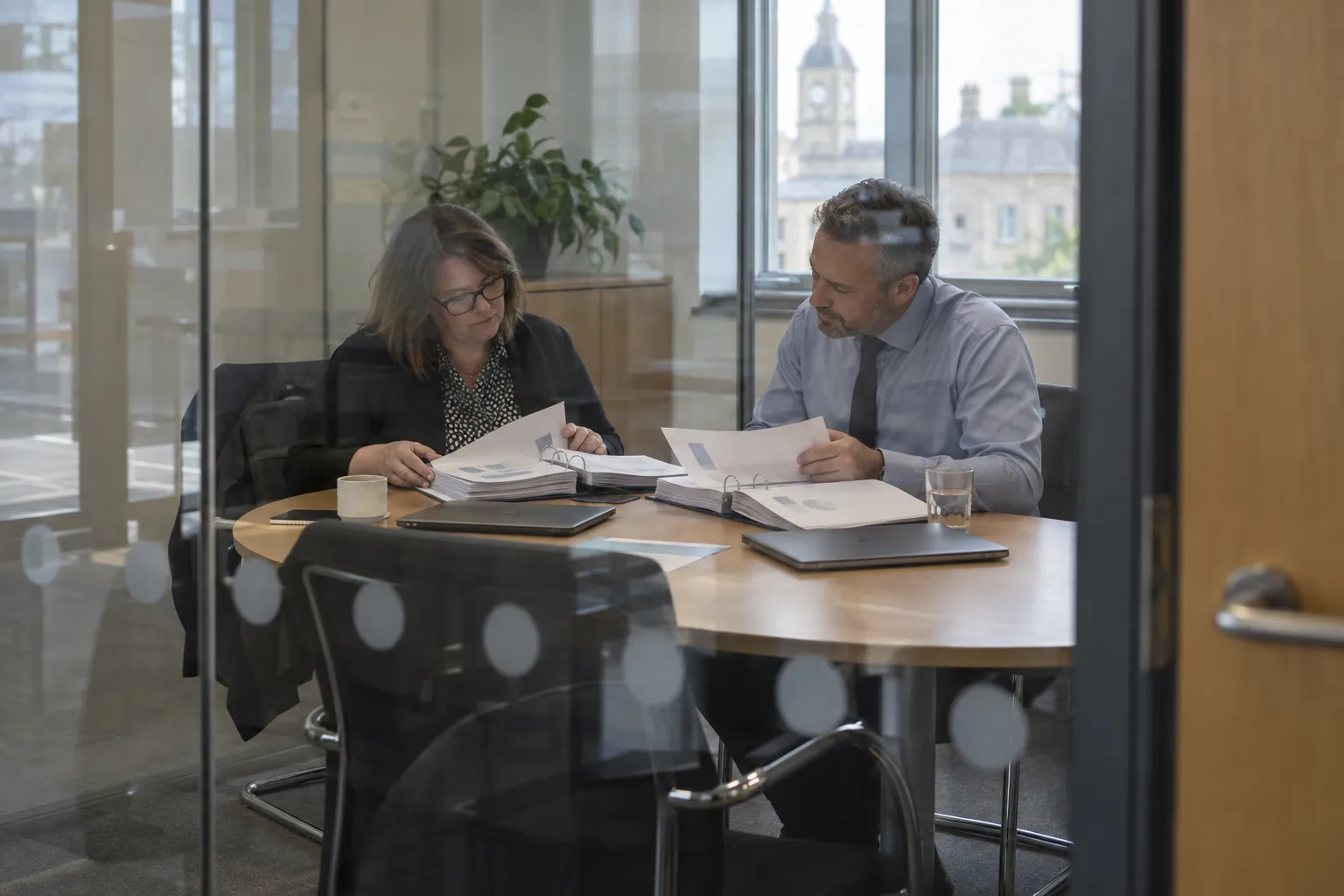 Two officers comparing tender documents in a glass-walled meeting room.