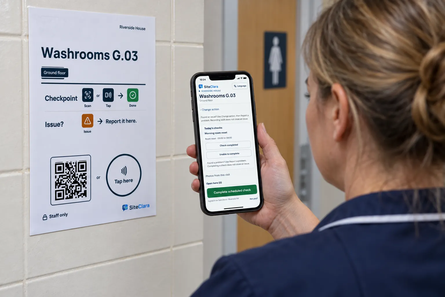 A supervisor checking a cleaning app on a phone beside a checkpoint poster on a washroom wall.